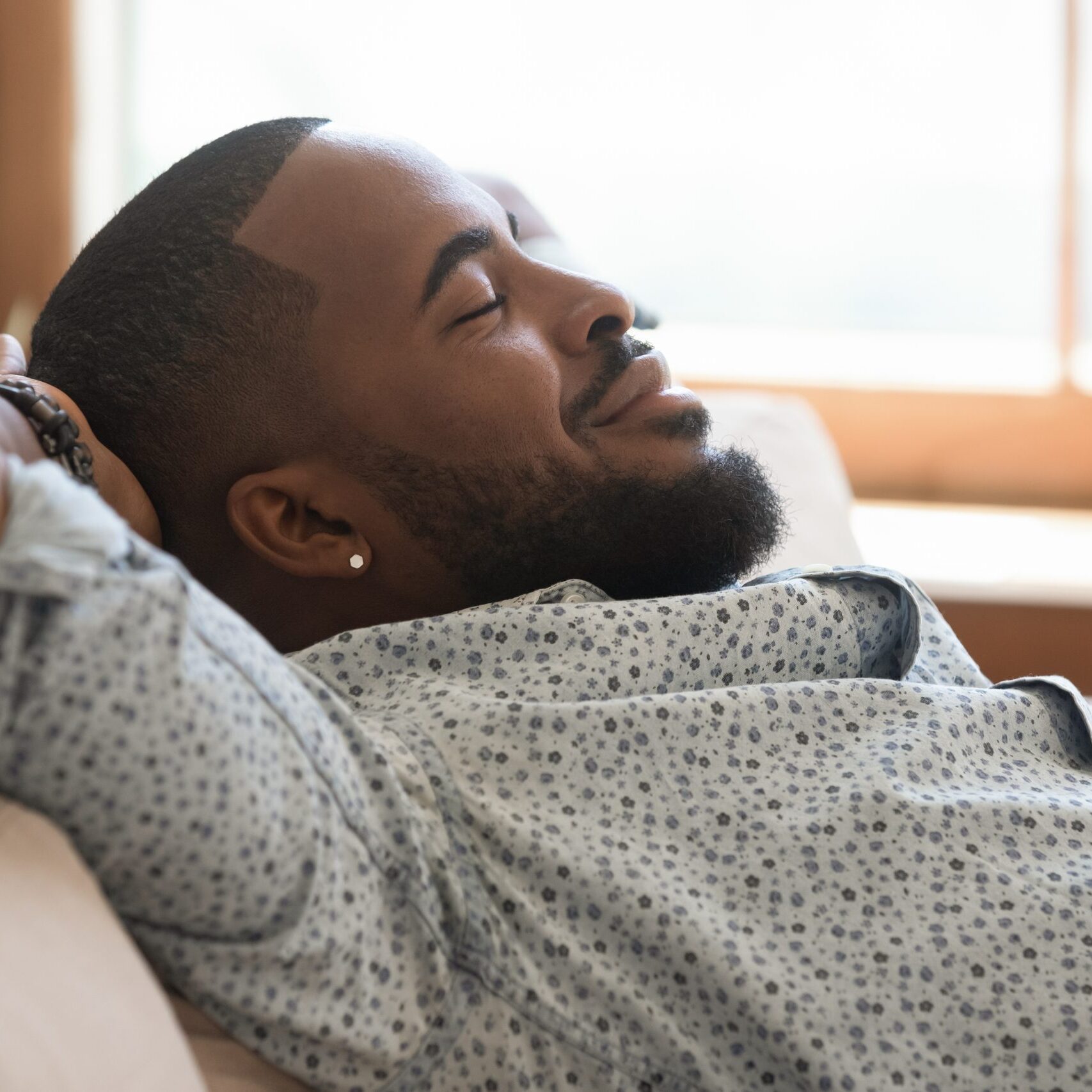 Calm lazy african american young man relaxing leaning on sofa at home, happy mindful guy holding hands behind head breathing fresh air lounge on couch enjoy stress free peaceful day with eyes closed